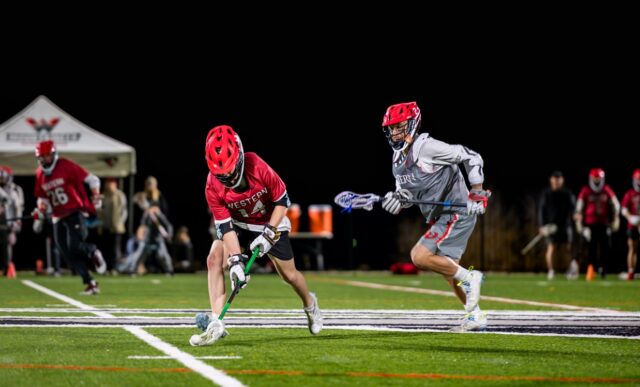 Two lacrosse athletes run on a field at night.