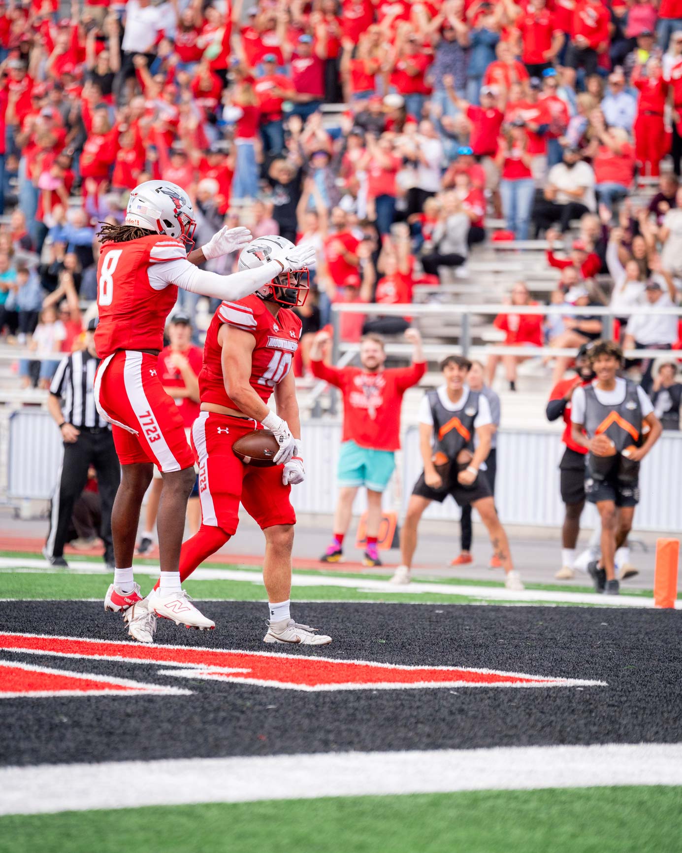 Western player #10 celebrates in the endzone after completing a touchdown his teammate rushed next to him to celebrate by slaping the top of #9s helmet. The stands are in the background and you can see fans standing and cheering, as well as the people on the sidlines.