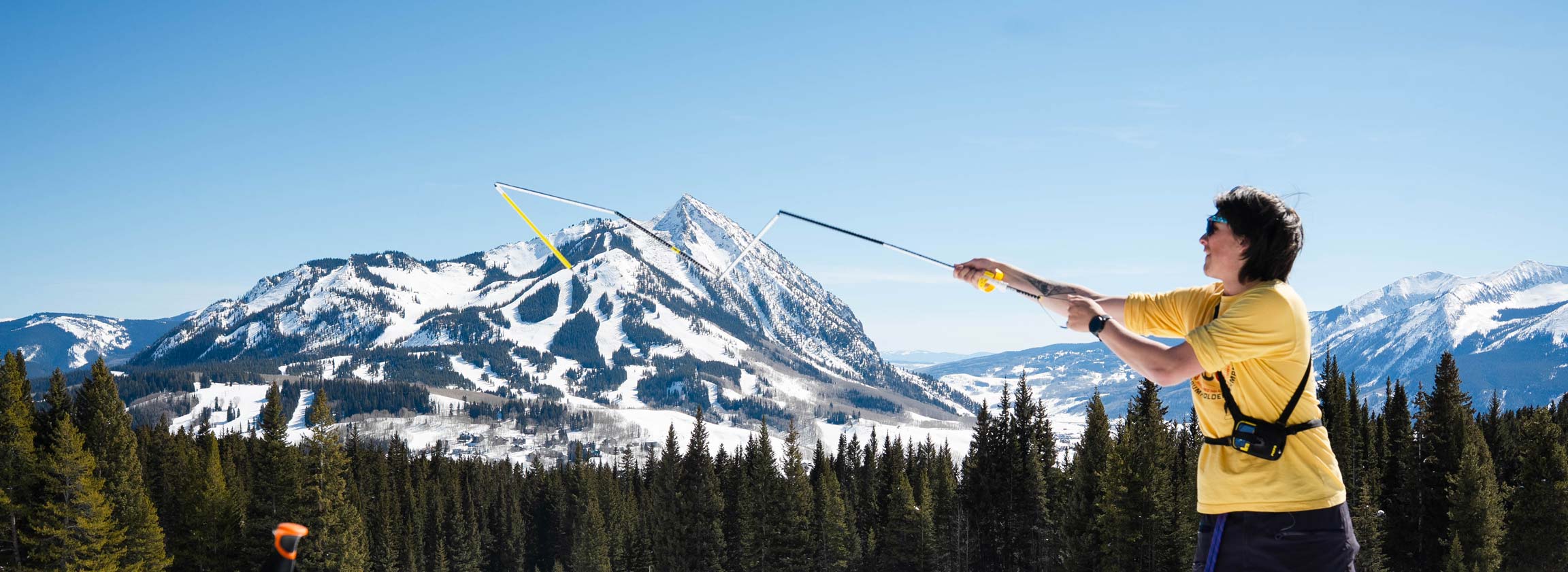 A person in a yellow shirt stands on a snowy slope, holding ski poles and looking toward a mountain range under a clear blue sky.