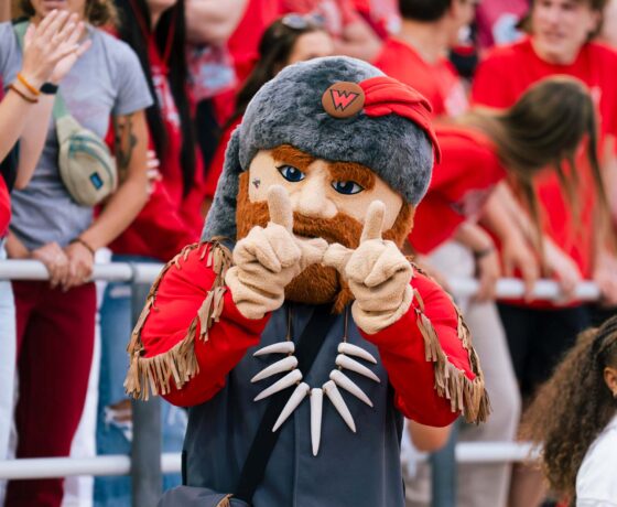 Mad Jack holds his hands up in the shape of a W while in the stands at a football game in Mountaineer Bowl.