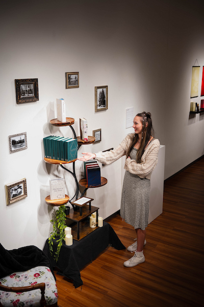 A person gestures toward a multi-tiered bookshelf displaying various books and framed photographs in a warmly lit indoor space.