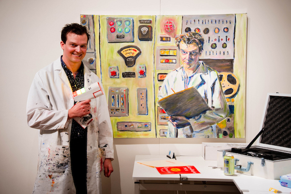 An individual in a paint-splattered lab coat stands beside a large painting depicting a scientist in a lab setting, with art supplies and equipment visible nearby.