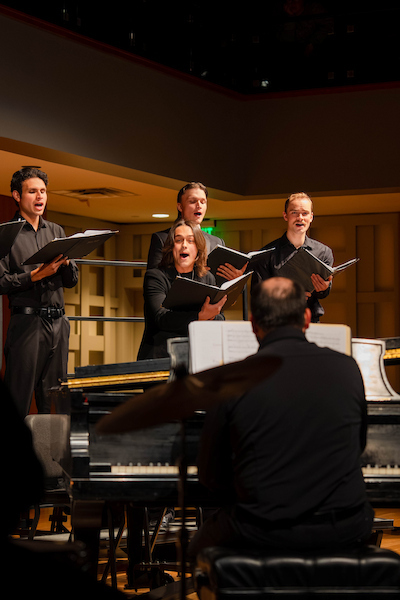 A group of four singers in black attire performs on stage with a pianist.