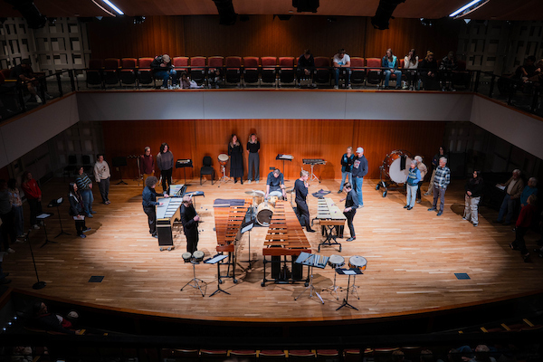 A group of musicians plays a variety of percussion instruments on a wooden stage in a concert hall with an audience seated in the balcony.