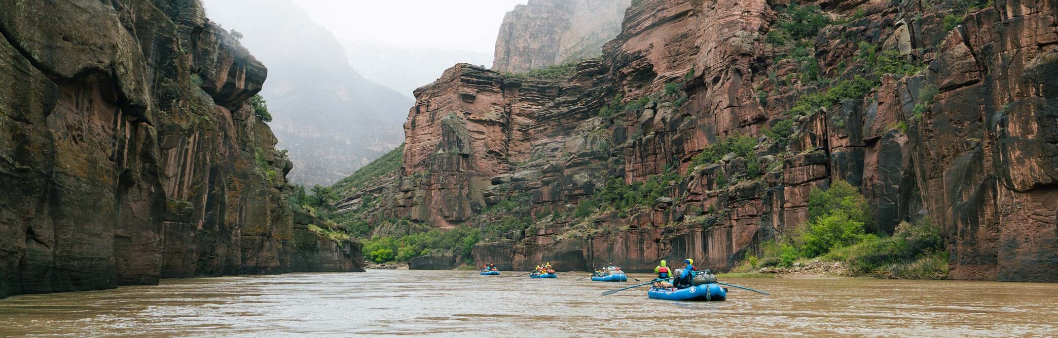 Rafters navigate a muddy river through a narrow canyon flanked by towering red rock cliffs under an overcast sky.
