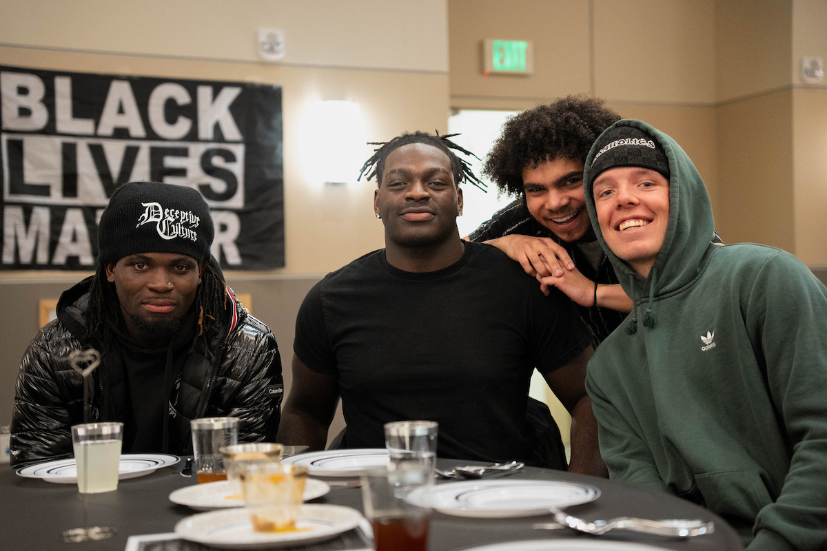 Four individuals sit together at a dining table in a room with a "Black Lives Matter" banner, smiling and posing for the camera.