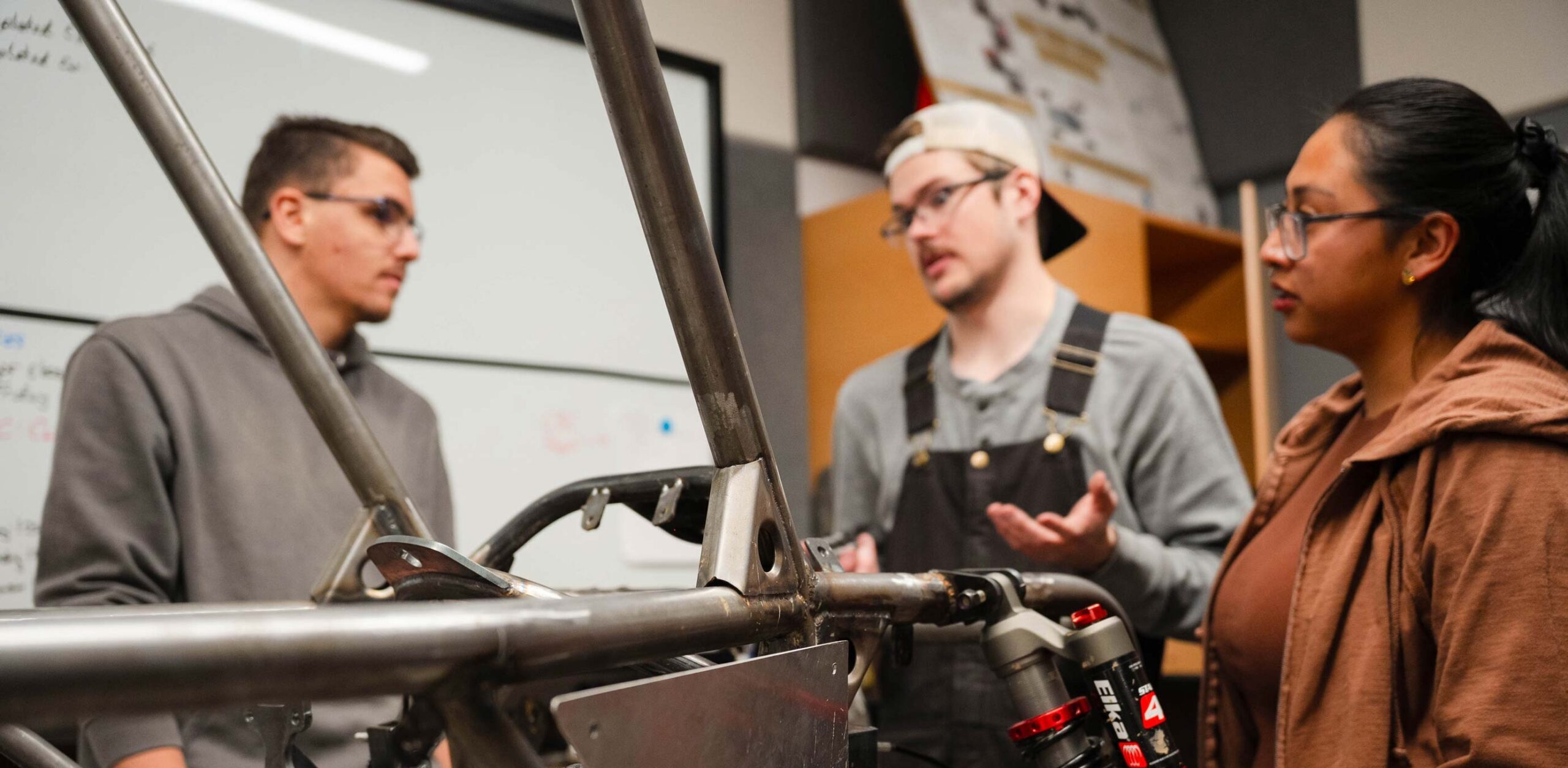 Three individuals examine a metal vehicle frame in a workshop setting.