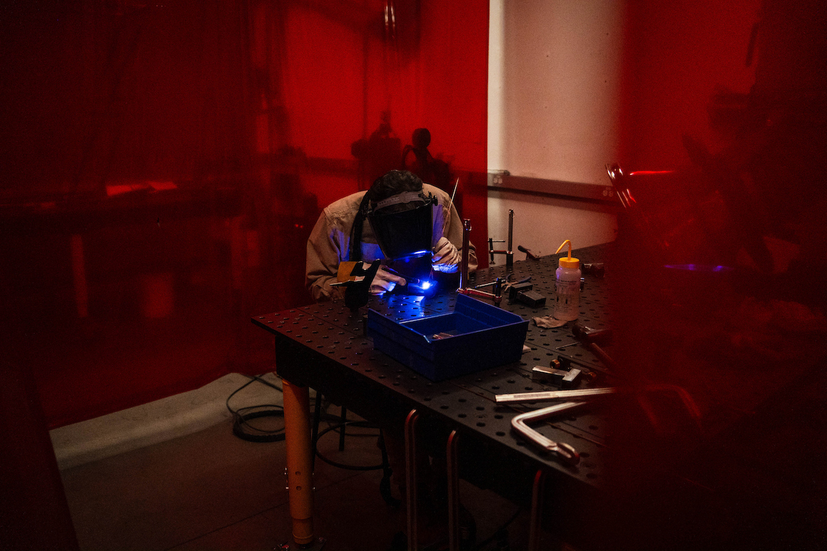 A person wearing a welding helmet works on a metal piece under a bright blue light, surrounded by tools and equipment in a dimly lit workshop bathed in red ambient light.