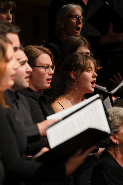 A choir performs with expressive faces, holding music sheets and singing in unison.