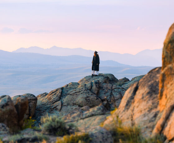 A person in a graduation gown stands atop rocky terrain.