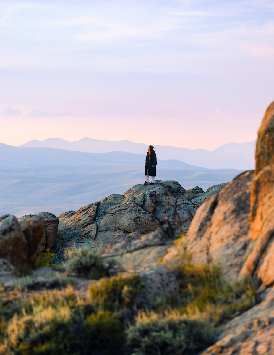 A person in a graduation gown stands atop rocky terrain.