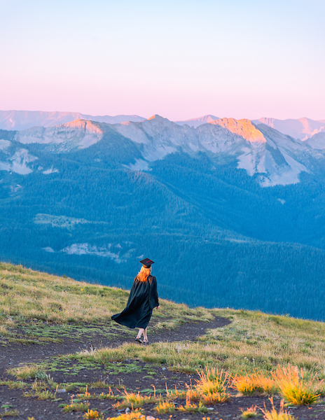 A student wearing a cap and gown walks down a high alpine ridgeline at sunrise.