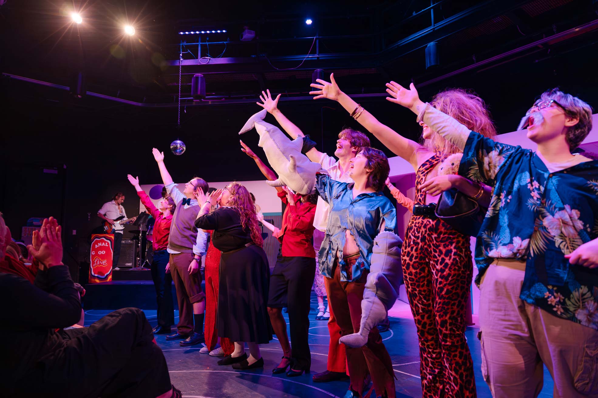 A group of performers on stage, arms raised in celebration, illuminated by vibrant stage lights with a band playing in the background.