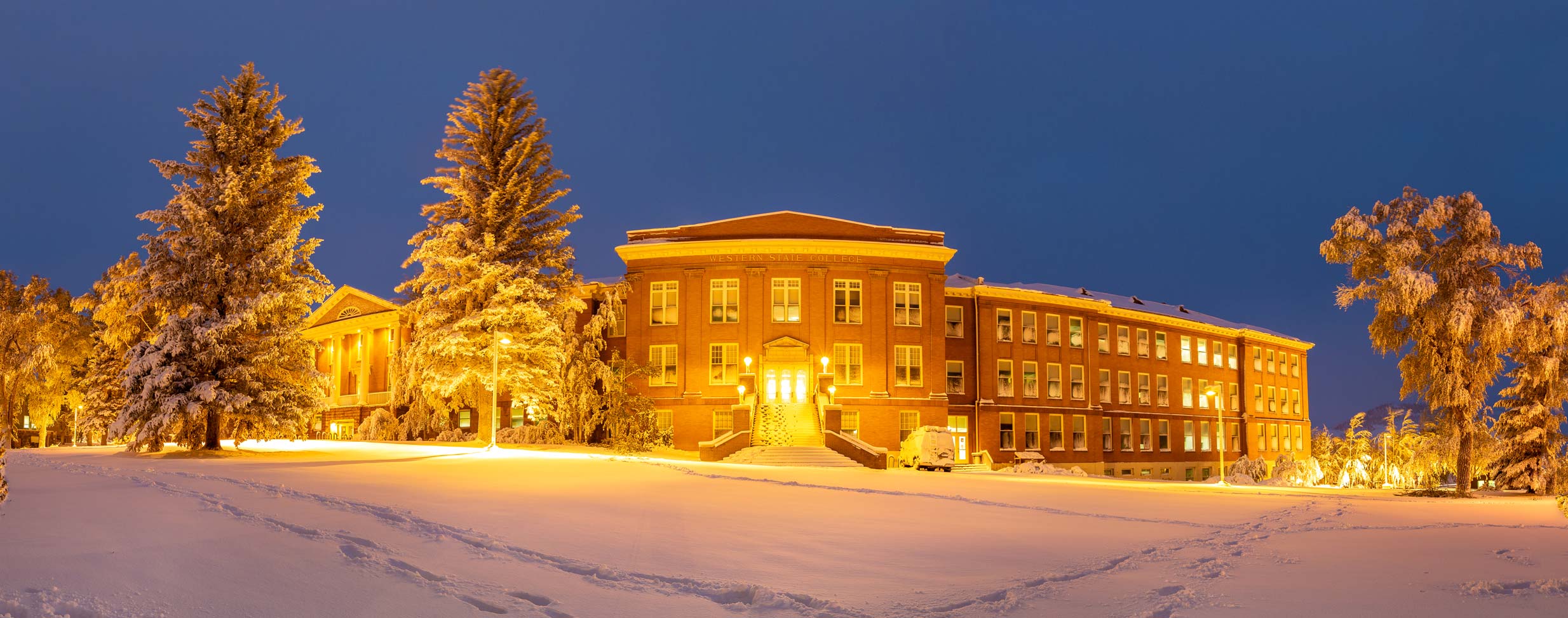 A warmly lit brick building stands amidst snow-covered trees and a snowy foreground under a deep blue twilight sky.