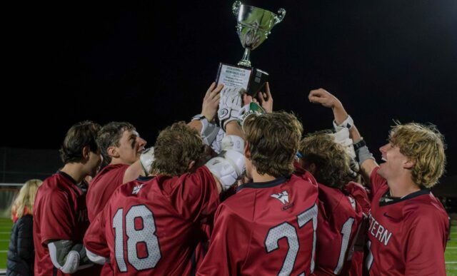 A group of Western Mens Lacrosse players celebrate winning the alumni game. They are gathered in a group as they all hold up the Dan Johnson Memorial Cup trophy.