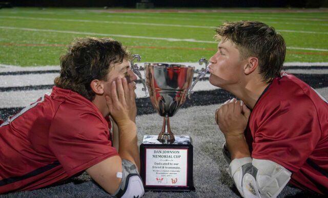 Two mens lacrosse players lay playfully on the ground on either side of the trophy, They both lean in to give the trophy a kiss.