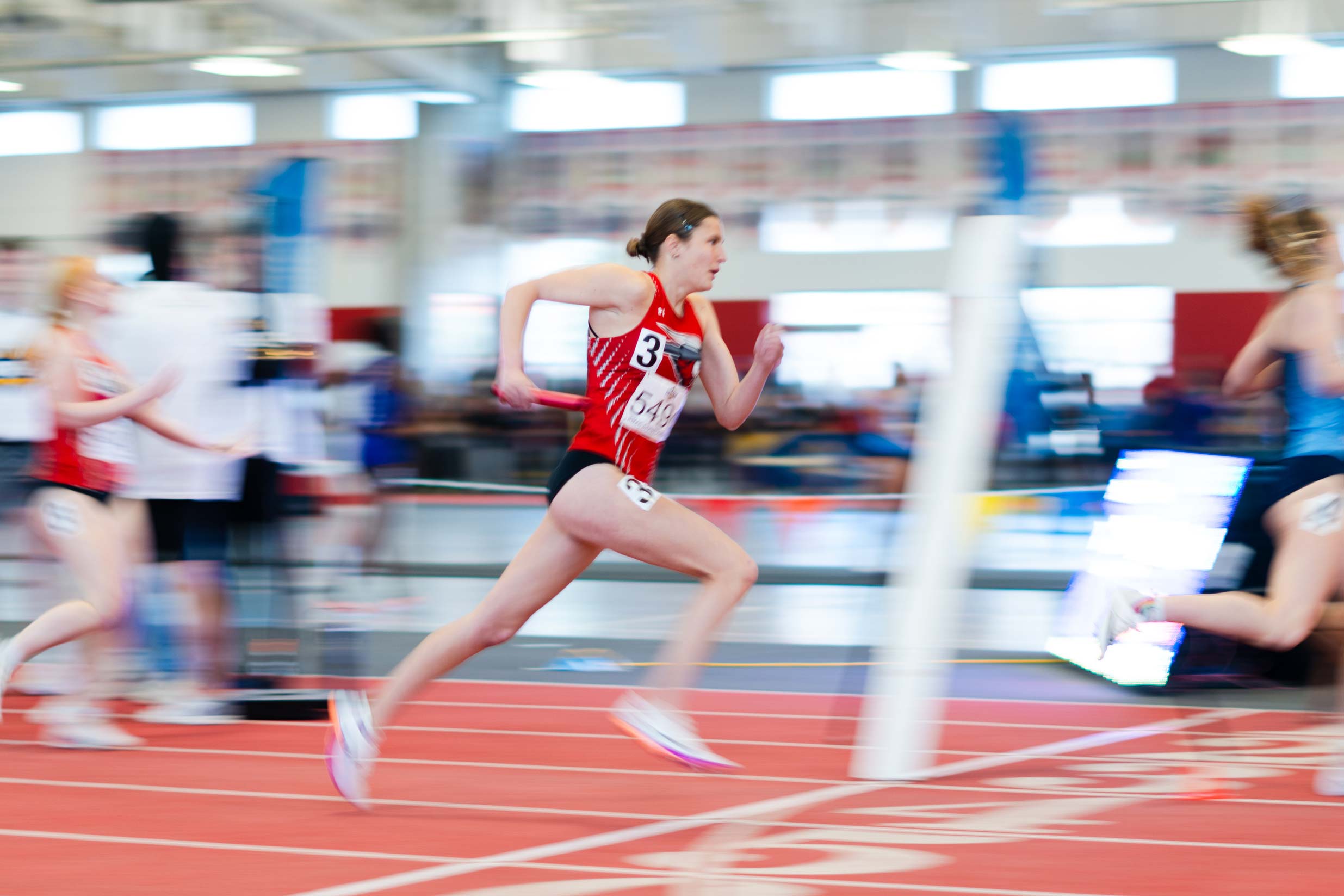 An athlete in a red uniform sprints on an indoor track during a race, with blurred competitors and surroundings emphasizing motion.