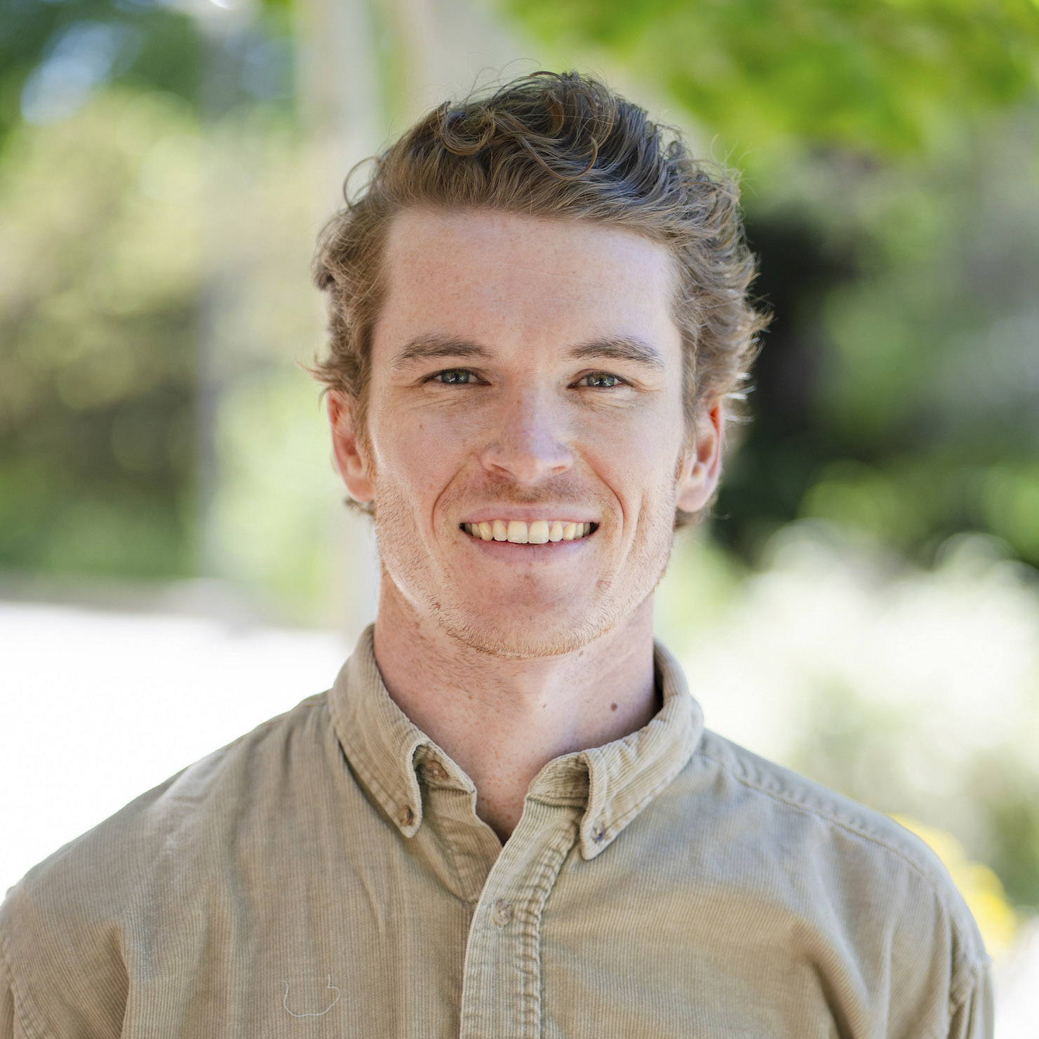 A smiling person with curly hair wears a beige corduroy shirt outdoors against a blurred green background.
