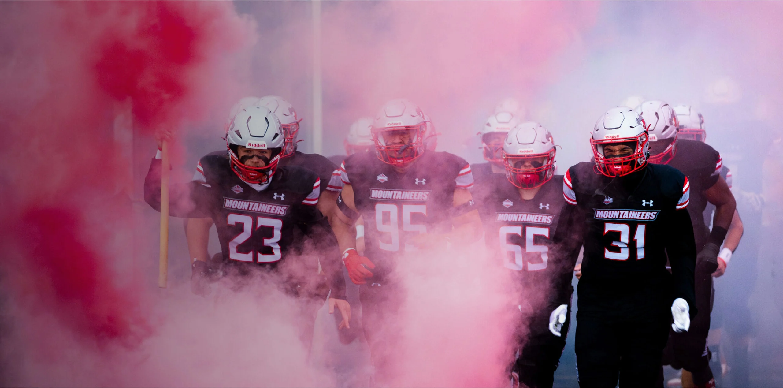 Football athletes run onto the field through pink smoke.